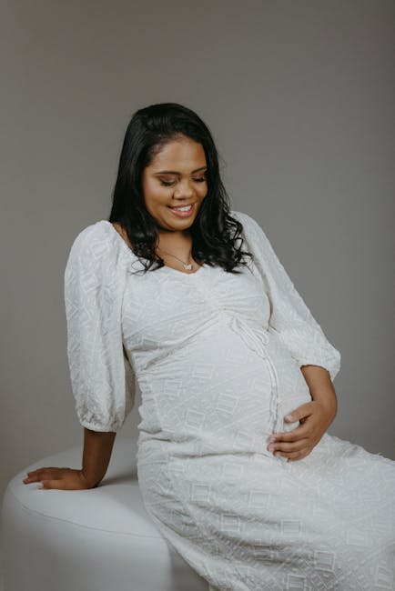A pregnant woman with dark, shoulder-length hair is sitting on a white cushioned surface against a plain, light grey background. She is wearing a white, textured, long-sleeved dress that fits snugly over her baby bump. Her left hand is gently resting on her stomach, while her right hand is positioned on the cushion beside her for support. She is smiling softly and looking slightly downward, conveying a sense of calm and contentment. The setting appears to be a studio or indoor environment with soft, even lighting highlighting her facial features and the fabric details of her dress. Although the image focuses on her, it can be associated with themes of home and family, which align with moving and relocation services such as those offered by Man with Van Lewisham, especially when considering preparations for a new home during a move.