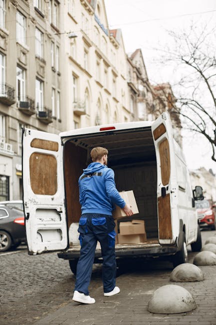 A man with a beard and wearing a black cap, white polo shirt, and jeans is kneeling on the pavement next to an open cardboard box, using a marker to label the box during a home relocation process. Behind him, there are multiple stacked cardboard boxes, some partially open, containing packing materials such as plastic wrap and paper. The boxes are arranged nearby, ready for loading into a van, which is visible with its open side door revealing the interior. The setting appears to be outside a property, with the van parked close to the entrance on a paved surface, under natural daylight. This scene captures a stage of packing and furniture transport during a professional removals service, as provided by Man with Van Lewisham, supporting efficient packing and loading for a house removal.