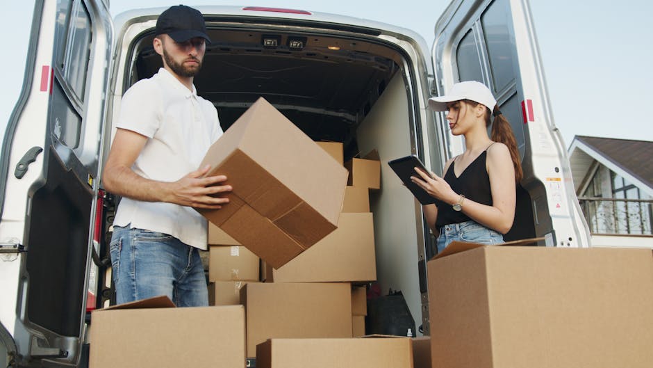 A male removalist from Man with Van Lewisham, wearing a white t-shirt and blue work overalls, is seated on the edge of a white van with its sliding door open. He is carefully handling a cardboard box,