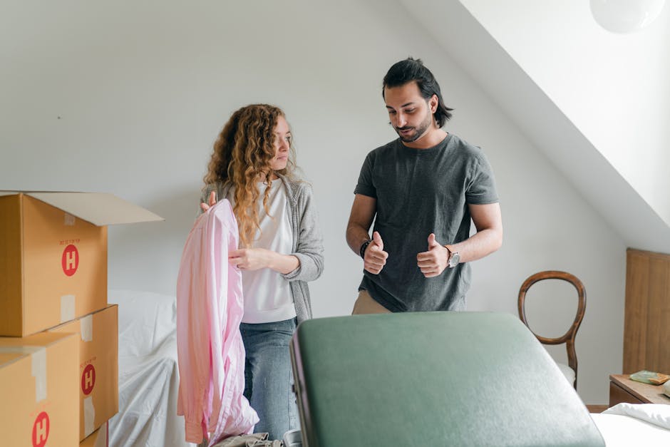 A man and a woman inside a residential room preparing for a home relocation, with several cardboard moving boxes stacked on the left side, some open and labeled with a red 'H' symbol. The woman, with