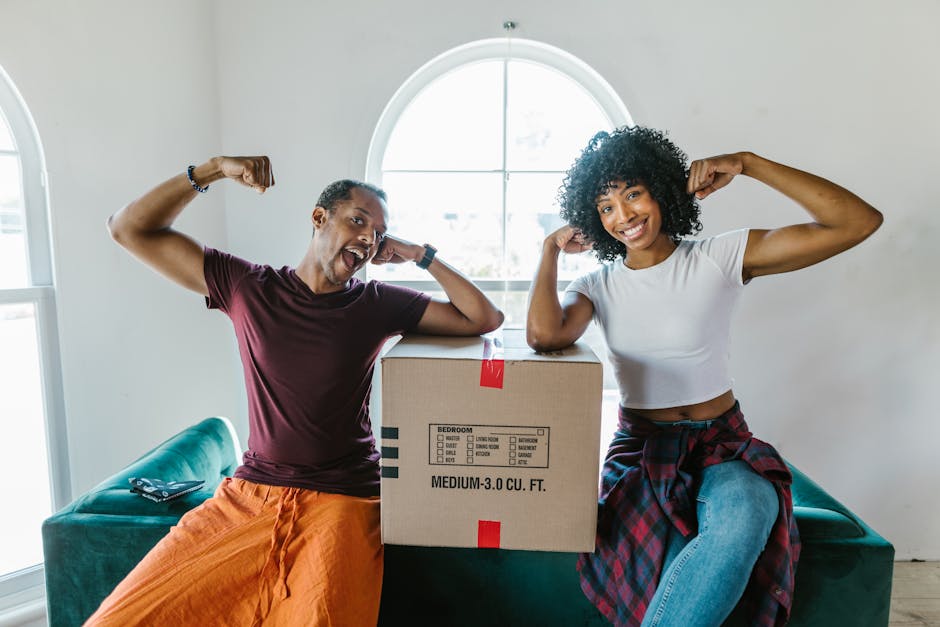 A man and a woman inside a residential room preparing for a home relocation, with several cardboard moving boxes stacked on the left side, some open and labeled with a red 'H' symbol. The woman, with