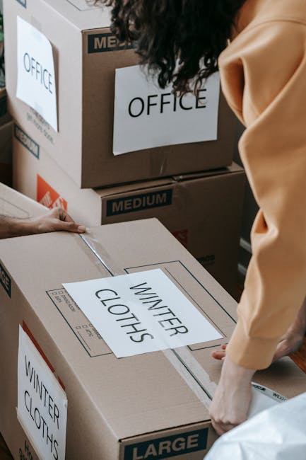 A person wearing a beige coat is handling a large cardboard box labeled 'WINTER CLOTHES' during a home relocation or packing process, surrounded by several other labeled boxes marked 'OFFICE' and 'MED