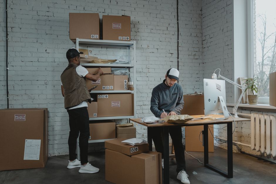A person wearing a beige coat is handling a large cardboard box labeled 'WINTER CLOTHES' during a home relocation or packing process, surrounded by several other labeled boxes marked 'OFFICE' and 'MED