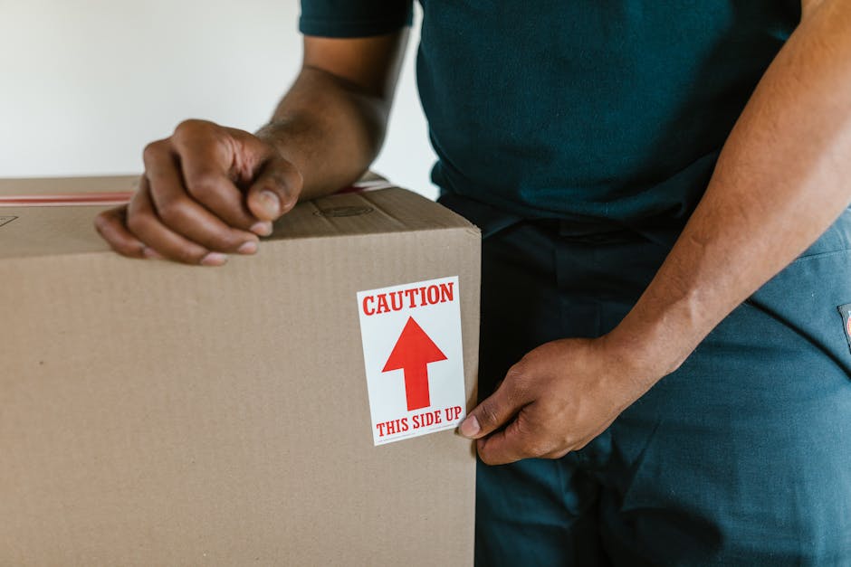 Close-up of a person wearing a dark green shirt and blue trousers, gripping a large cardboard box with a red and white CAUTION label indicating 'THIS SIDE UP' and an upward arrow. The individual is in