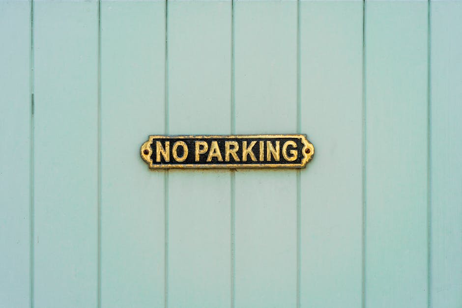 A close-up view of a rectangular metal 'No Parking' sign with gold lettering and a decorative border, mounted on a light green wooden wall composed of vertical panels. The sign is centrally positioned